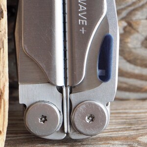 May include: Close-up of a silver multi-tool with the text "WAVE+" visible. The tool has a blue accent and is set against a wooden background. The tool's rivets are visible.
