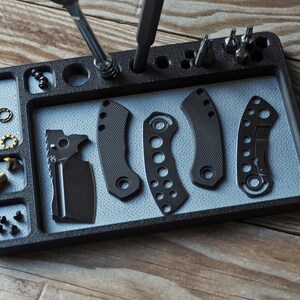 May include: A black organizer tray with various knife parts and tools. The tray has compartments for screws, washers, and other small components. Several knife handles and a blade are displayed.