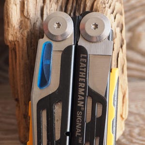 Könnte beinhalten: Nahaufnahme eines Leatherman Signal Multi-Tools. Das Werkzeug hat eine silberne Klinge, einen schwarzen Griff und gelbe Akzente. Der Text "LEATHERMAN SIGNAL" ist auf dem Griff aufgedruckt. Das Werkzeug ist teilweise geöffnet und zeigt seine Komponenten.