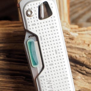 May include: Close-up of a silver multi-tool with a perforated design. The tool features a light green, rectangular accent and the word "MAN" is visible on the side. The tool is set against a wooden background.