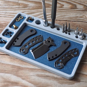 May include: A gray and blue parts tray with various tools and hardware. The tray features a blue textured surface and compartments holding screws, washers, and knife parts. Several screwdrivers and bits are also present, along with a small folding knife.
