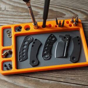 May include: An orange and gray tool tray with various tools and parts. The tray has compartments for small parts, and holes for holding screwdrivers and bits. Several black knife handles and a blade are also present.
