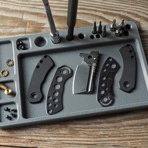 May include: A gray parts tray with various tools and components for knife assembly. The tray holds a knife blade, handle scales, screws, and a screwdriver. The components are arranged on a wooden surface.