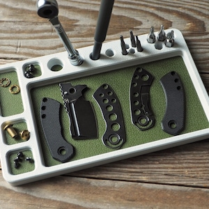 May include: A white rectangular tool organiser with a green interior. It holds various tools, including a ratchet, screwdriver, and several bits. The organiser also contains several black knife handles and small hardware pieces.