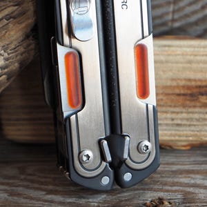 May include: Close-up of a multi-tool with a black handle and silver accents. The tool features two orange-colored inserts and a silver clip. The image is shot on a wooden surface.