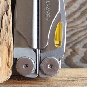 May include: Close-up of a silver multi-tool with the text "WAVE +" on the side. The tool has a yellow-tinted component and is partially open, revealing a blade. The background is a wooden surface.