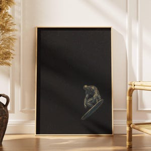 May include: Art print in a gold frame, depicting a surfer on a surfboard against a dark backdrop. The artwork is displayed in a room with neutral tones. The surfer is rendered in shades of grey and brown. The overall theme is surfing.