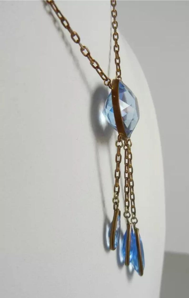 May include: A gold chain necklace with a large, faceted, light blue crystal pendant. The pendant is attached to three smaller, teardrop-shaped, light blue crystals.