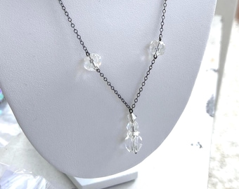 Peut inclure: Un collier délicat composé d'une fine chaîne en métal gris foncé, orné de deux perles en cristal transparent à facettes espacées le long de la chaîne, avec un pendentif central formé de deux perles en cristal transparent superposées.