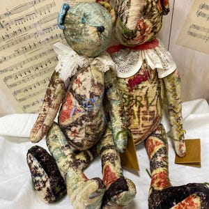 Puede incluir: Dos osos de peluche hechos a mano con tela de estilo vintage. Un oso tiene un lazo azul y un cuello de encaje blanco, el otro tiene un cuello rojo. Ambos tienen cuerpos estampados con colores apagados y est&aacute;n sentados sobre una superficie blanca.