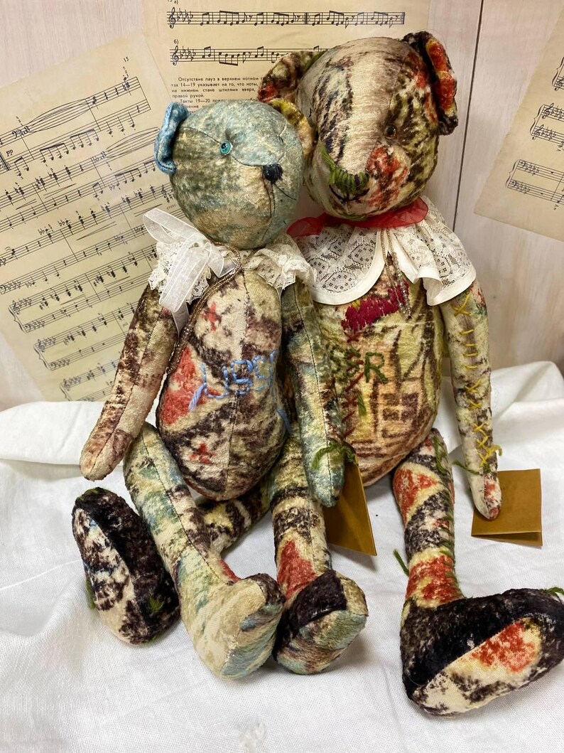 Puede incluir: Dos osos de peluche hechos a mano con tela de estilo vintage. Un oso tiene un lazo azul y un cuello de encaje blanco, el otro tiene un cuello rojo. Ambos tienen cuerpos estampados con colores apagados y est&aacute;n sentados sobre una superficie blanca.