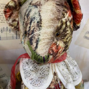 Puede incluir: Un oso de peluche de estilo vintage con un cuerpo de tela estampada en tonos marrones, verdes y rojos. El oso tiene un cuello de encaje blanco con una cinta roja y un peque&ntilde;o collar. La cara del oso tiene detalles cosidos.