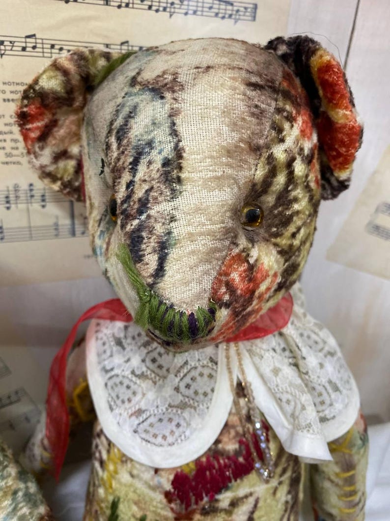 Puede incluir: Un oso de peluche de estilo vintage con un cuerpo de tela estampada en tonos marrones, verdes y rojos. El oso tiene un cuello de encaje blanco con una cinta roja y un peque&ntilde;o collar. La cara del oso tiene detalles cosidos.