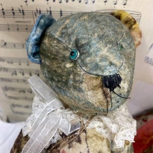 Puede incluir: Un oso de peluche de estilo vintage con una cabeza estampada en azul y verde y un cuello de encaje blanco. El oso tiene ojos turquesa y una nariz cosida negra. El cuerpo del oso est&aacute; estampado con dise&ntilde;os florales y abstractos.