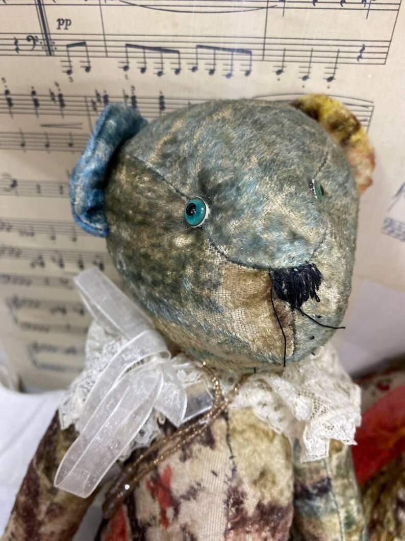 Puede incluir: Un oso de peluche de estilo vintage con una cabeza estampada en azul y verde y un cuello de encaje blanco. El oso tiene ojos turquesa y una nariz cosida negra. El cuerpo del oso est&aacute; estampado con dise&ntilde;os florales y abstractos.