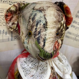 Puede incluir: Un oso de peluche de estilo vintage con cabeza y cuerpo de tela estampada. El oso tiene ojos de bot&oacute;n, una cinta roja y un cuello de encaje blanco. La tela presenta una mezcla de colores, incluyendo marr&oacute;n, rojo y verde.