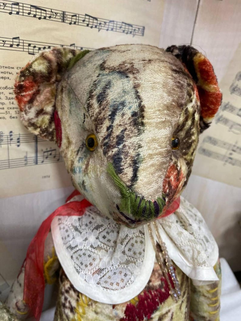 Puede incluir: Un oso de peluche de estilo vintage con cabeza y cuerpo de tela estampada. El oso tiene ojos de bot&oacute;n, una cinta roja y un cuello de encaje blanco. La tela presenta una mezcla de colores, incluyendo marr&oacute;n, rojo y verde.