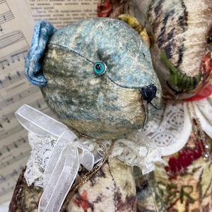 Puede incluir: Dos osos de peluche de estilo antiguo. Uno tiene una cabeza azul y dorada con una oreja azul y un ojo turquesa. El otro oso tiene una cabeza estampada con un cuello rojo. Ambos osos tienen cuellos de encaje y lazos.