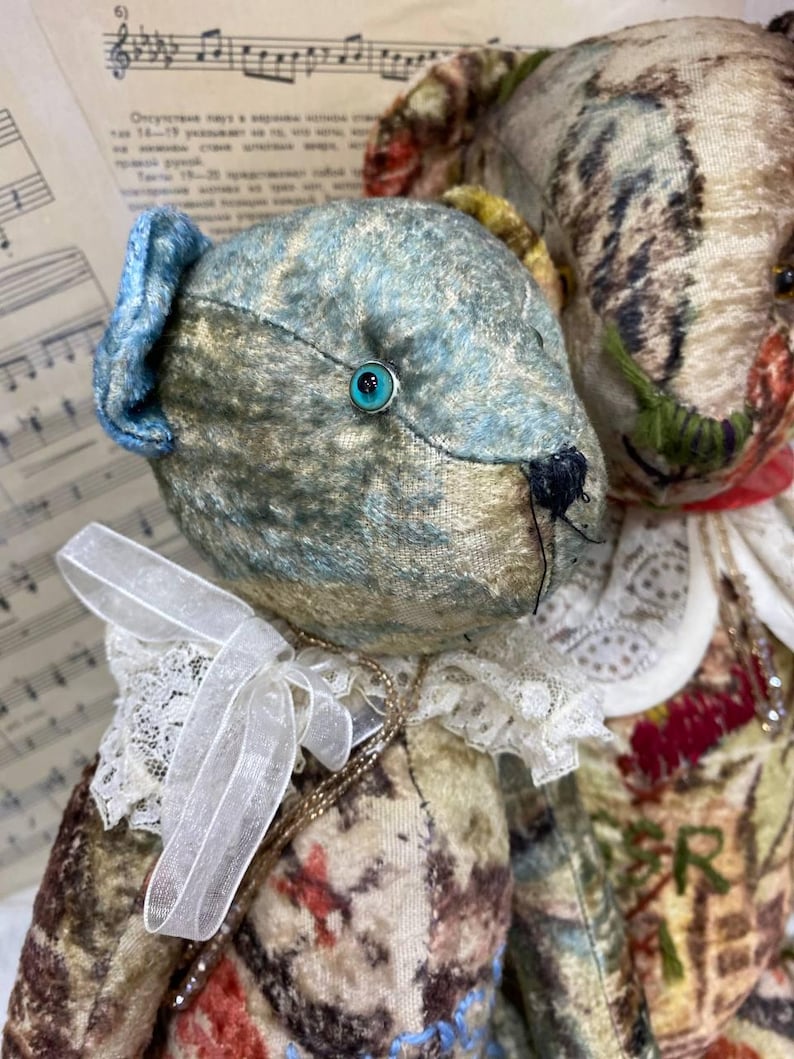 Puede incluir: Dos osos de peluche de estilo antiguo. Uno tiene una cabeza azul y dorada con una oreja azul y un ojo turquesa. El otro oso tiene una cabeza estampada con un cuello rojo. Ambos osos tienen cuellos de encaje y lazos.