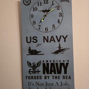 May include: A rectangular wall clock with a light blue background featuring a clock face, the words "US NAVY", and various navy-related graphics. The clock also includes the text "AMERICA'S NAVY FORGED BY THE SEA It's Not Just A Job, It's An Adventure!"