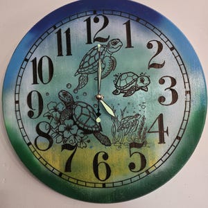 May include: Round wall clock with a sea turtle theme. The clock face features a gradient of blue and green, with black numerals and illustrations of turtles. The clock hands are silver and glow in the dark.