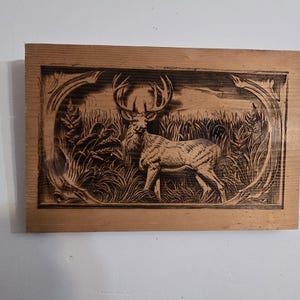 Puede incluir: Placa rectangular de madera con un detallado relieve de un ciervo con grandes astas en hierba alta. La obra de arte está enmarcada por un borde ornamentado, mostrando un tema rústico y natural.