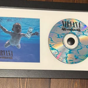 May include: Framed Nirvana "Nevermind" album art and CD. The artwork features a baby swimming underwater reaching for a dollar bill. The CD has a holographic surface with the band's name and album title.