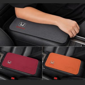 May include: A rectangular car center console armrest cover made of smooth material with stitched edges. It features the Honda logo in silver and red, along with a subtle embossed car outline. Available in gray, dark red, and orange color options.
