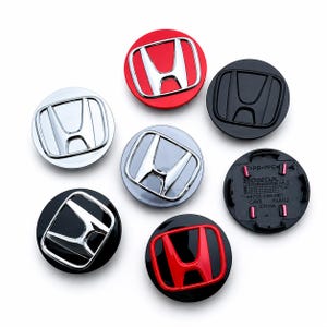 Può includere: Una collezione di sette coprimozzo circolari Honda in varie finiture, tra cui cromo, nero opaco, rosso e nero con emblemi cromati. Un tappo è capovolto per rivelare il lato inferiore in plastica nera con testo e segni stampati.