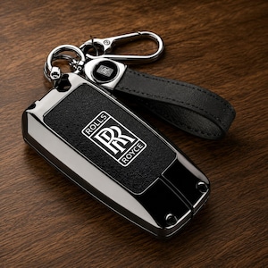 May include: A black and silver Rolls-Royce key fob featuring a textured black center panel with the white Rolls-Royce logo and text. The key fob is attached to a black leather loop and a silver metal carabiner clip, creating a complete key accessory.