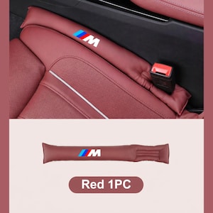 May include: A red car seat gap filler made of smooth material featuring the M logo with blue, purple, and red stripes. The long, cylindrical cushion is designed to fit between the seat and center console, shown installed in a vehicle interior.