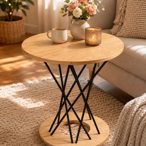 May include: A round side table with a light wood top and base, connected by a series of thin, black metal rods arranged in a crisscross, hourglass-shaped pattern. The table surface holds a ceramic mug, a small vase with flowers, and a glass candle.
