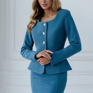 May include: A teal two-piece suit featuring a fitted jacket with a square neckline, three white buttons, and a peplum waist. The matching pencil skirt completes the professional ensemble. The text "Shari Ja" is visible at the bottom.