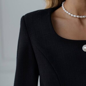 May include: Close-up of a black tweed jacket with a square neckline and a pearl button. A pearl necklace is visible. The jacket has a textured weave and a tailored fit, suggesting a classic style. The image focuses on the upper body and neckline.