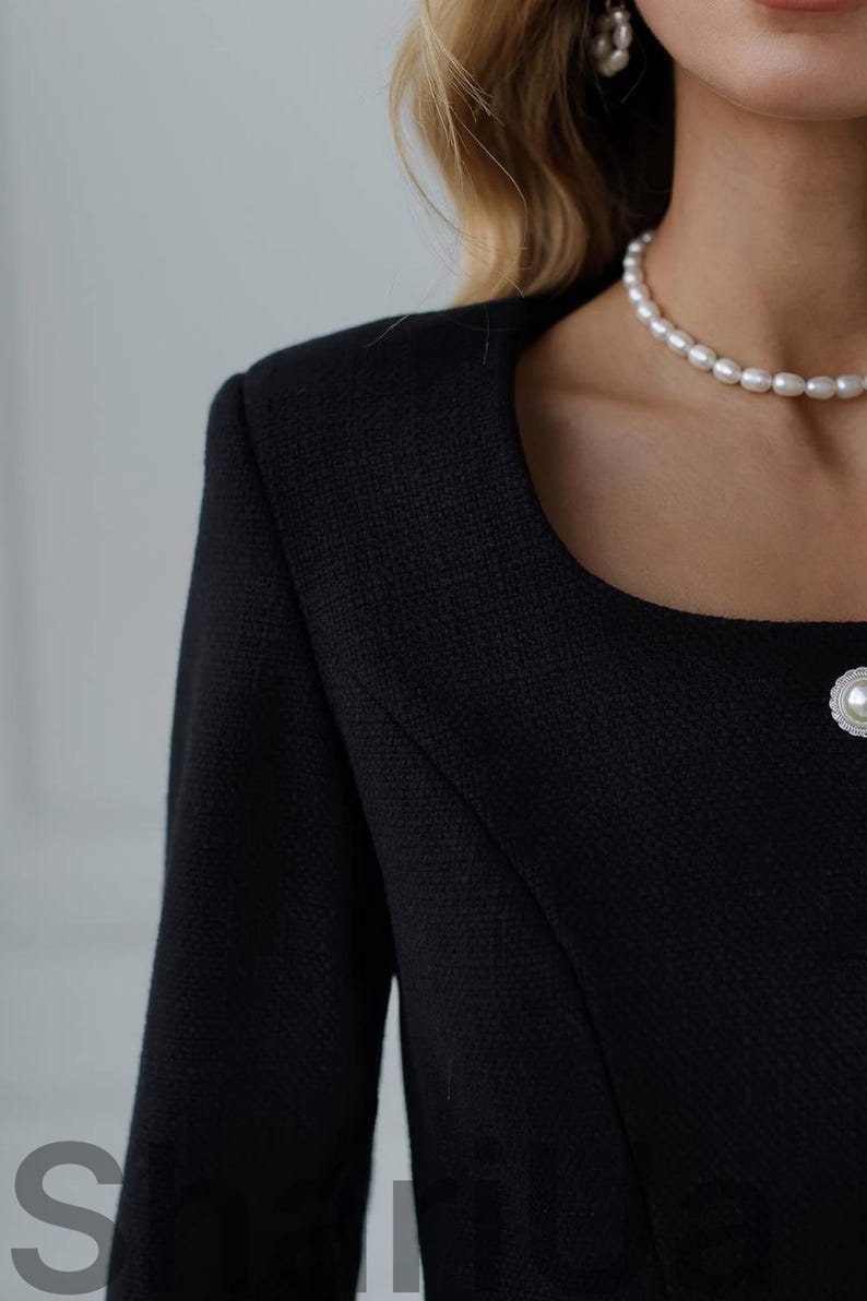 May include: Close-up of a black tweed jacket with a square neckline and a pearl button. A pearl necklace is visible. The jacket has a textured weave and a tailored fit, suggesting a classic style. The image focuses on the upper body and neckline.