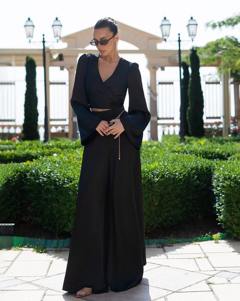 May include: A black two-piece outfit featuring a wrap-style top with long, flared sleeves and high-waisted, wide-leg pants. A gold chain belt accents the waist. The outfit is made of a flowing fabric, suitable for a formal or semi-formal occasion.