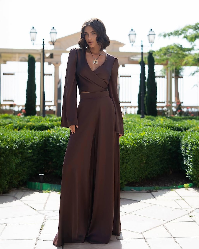 May include: A brown two-piece outfit featuring a wrap-style long-sleeve top and wide-leg pants. The top has a V-neck and flared sleeves. The outfit is made of a flowing fabric and is suitable for formal occasions.