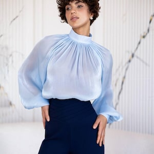 May include: A pale blue blouse with a high neck and long sleeves, worn with navy blue high-waisted trousers. The blouse features a gathered neckline, and the trousers have a pleated design. The outfit is elegant and stylish.