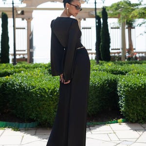 May include: A long, black jumpsuit with wide legs and long, flared sleeves. A gold-colored chain belt cinches the waist. The outfit is worn outdoors in front of a manicured green hedge and a stone archway.