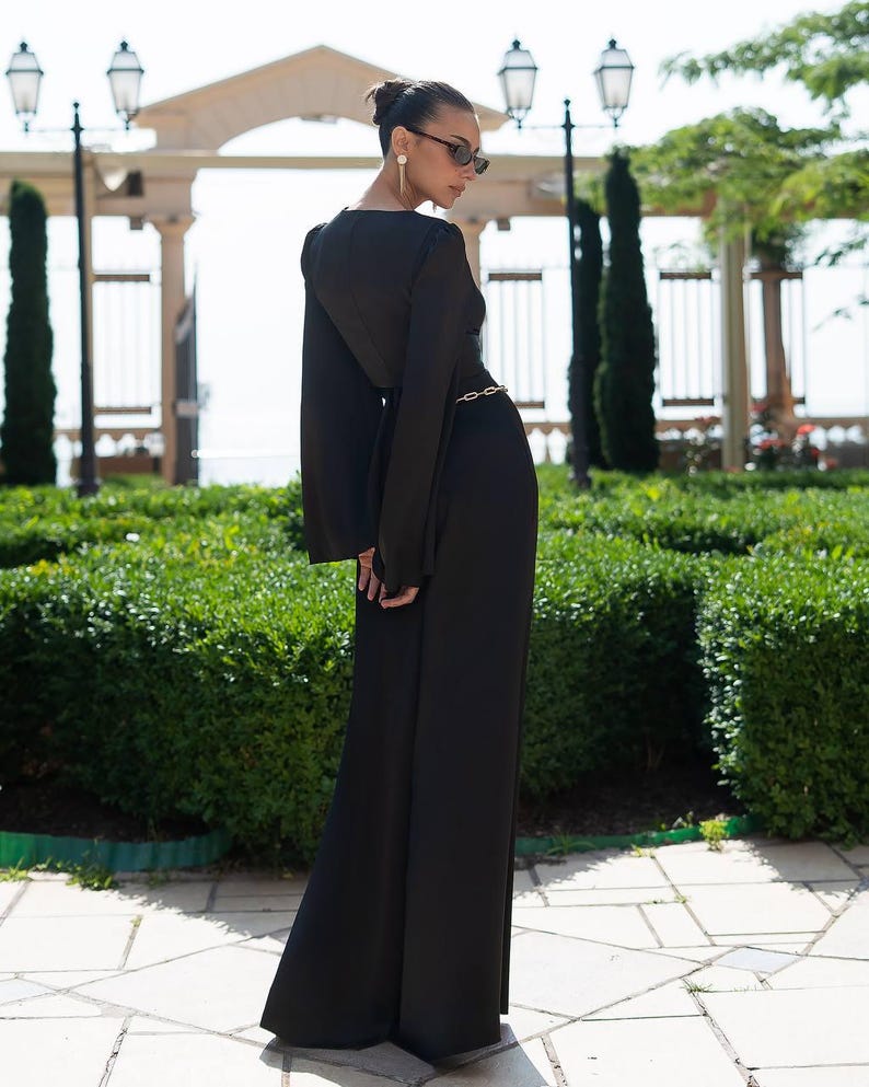 May include: A long, black jumpsuit with wide legs and long, flared sleeves. A gold-colored chain belt cinches the waist. The outfit is worn outdoors in front of a manicured green hedge and a stone archway.