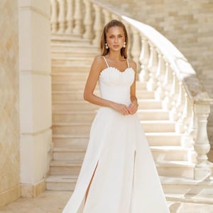 May include: A white midi dress with a sweetheart neckline and spaghetti straps. The dress features a high slit on each side and is worn by a person standing in front of a staircase with a white balustrade.