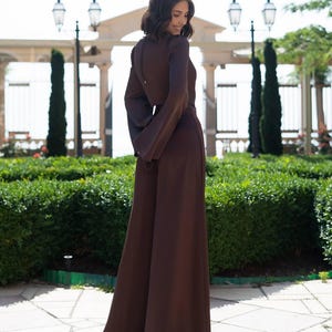 May include: A long-sleeved, brown jumpsuit with wide-leg pants. The jumpsuit features a keyhole back and a matching belt. The model is standing outside in front of a manicured hedge and a decorative gate.