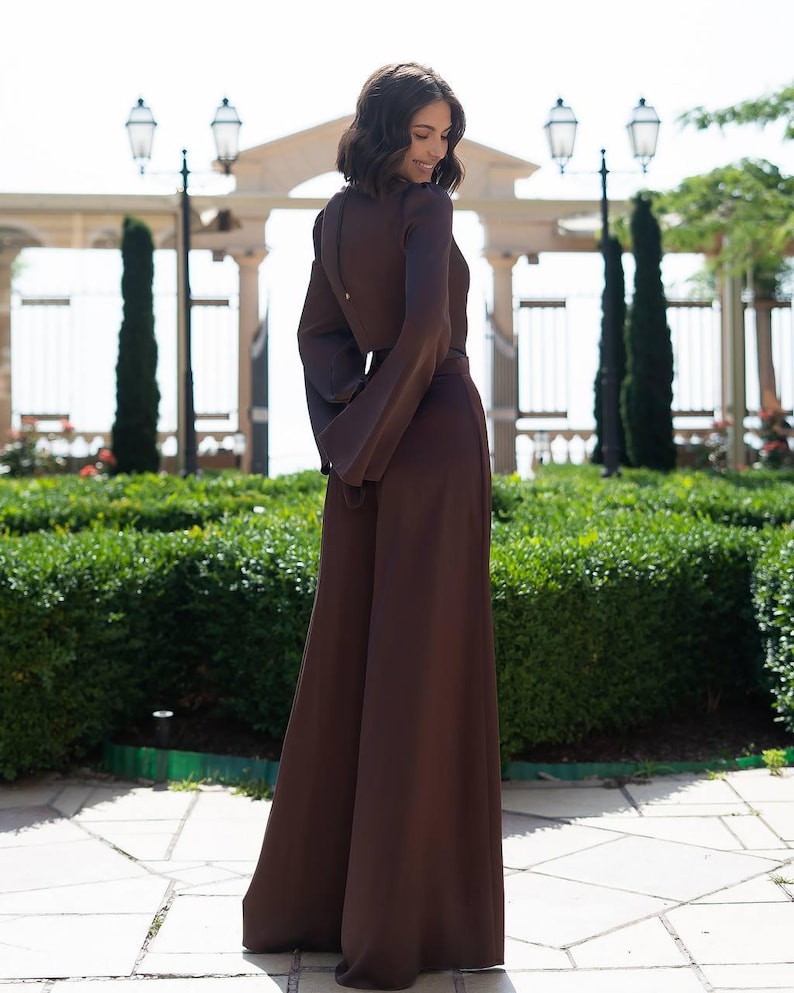 May include: A long-sleeved, brown jumpsuit with wide-leg pants. The jumpsuit features a keyhole back and a matching belt. The model is standing outside in front of a manicured hedge and a decorative gate.
