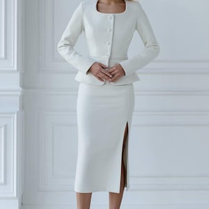 May include: A white two-piece suit featuring a fitted jacket with a square neckline and button closure, paired with a knee-length skirt with a side slit. The suit is made of a textured fabric. The text "ShariUa" is visible at the bottom.