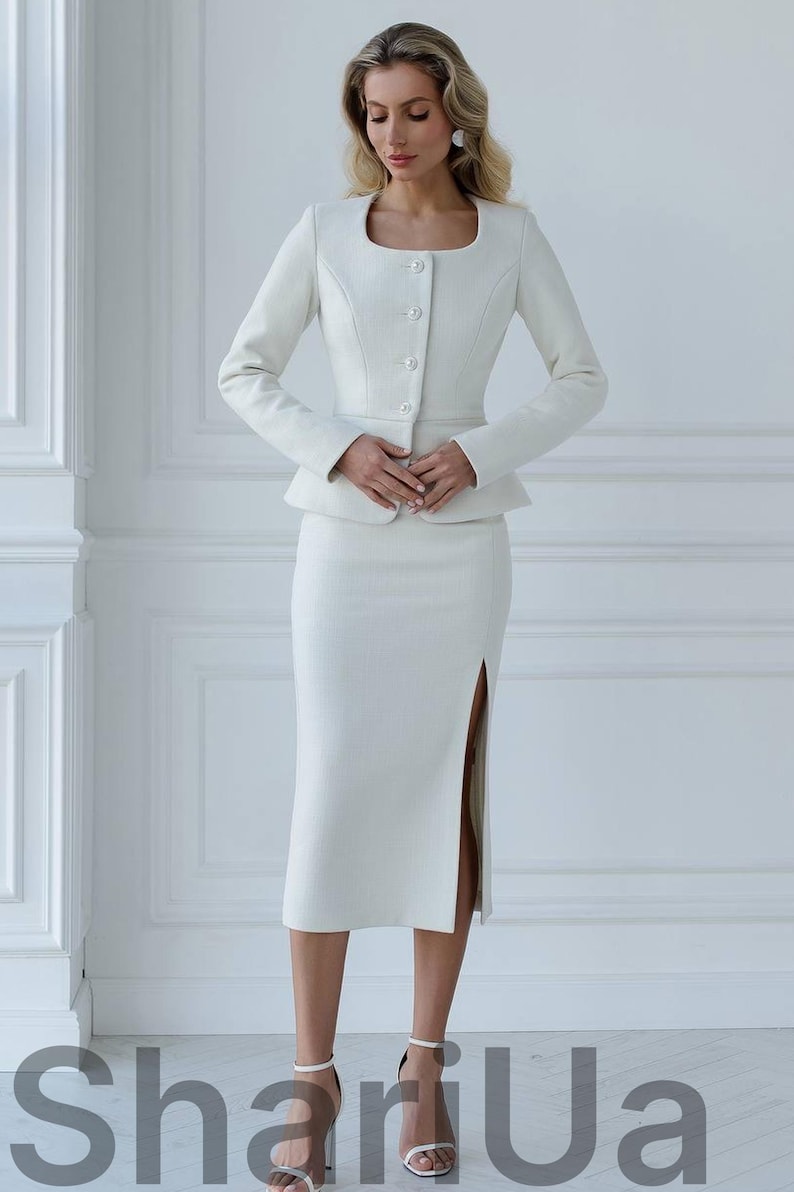May include: A white two-piece suit featuring a fitted jacket with a square neckline and button closure, paired with a knee-length skirt with a side slit. The suit is made of a textured fabric. The text "ShariUa" is visible at the bottom.