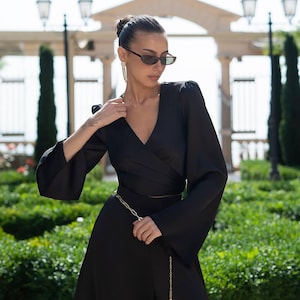 May include: A black wrap dress with long, flared sleeves and a gold chain belt. The dress has a V-neckline and a flowing skirt. The person is wearing sunglasses and earrings. The background features greenery and a stone structure.