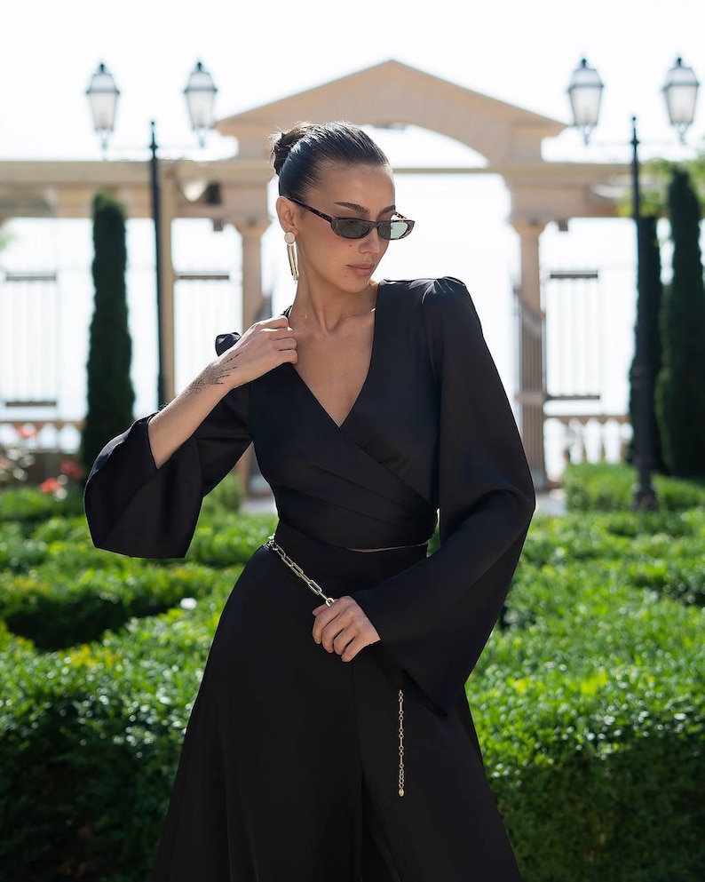May include: A black wrap dress with long, flared sleeves and a gold chain belt. The dress has a V-neckline and a flowing skirt. The person is wearing sunglasses and earrings. The background features greenery and a stone structure.