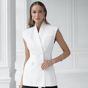 以下が含まれることがあります： ダブルブレストのデザインの白いノースリーブブレザー。 ブレザーは体に合ったフィット感で、大きなボタンが付いています。 モデルは黒いパンツと金のブレスレットを着用しています。 背景はプレーンな白い壁です。