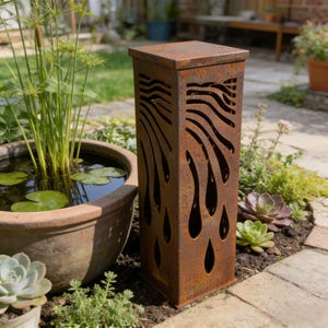 Könnte beinhalten: Eine verwitterte, rechteckige Gartenlaterne aus Metall mit quadratischer Oberseite. Die Seiten weisen ausgeschnittene Designs von fließendem Wasser oder Flammenformen auf. Die Laterne hat eine rostbraune Farbe und ist von Pflanzen umgeben.