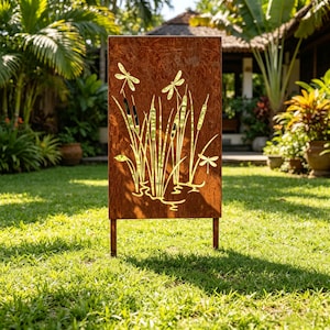 May include: A rectangular, weathered-look metal garden screen with a cut-out design of cattails and dragonflies. The screen is brown with a golden-yellow design and stands on two legs, placed on a green lawn. The background features lush greenery and a building.